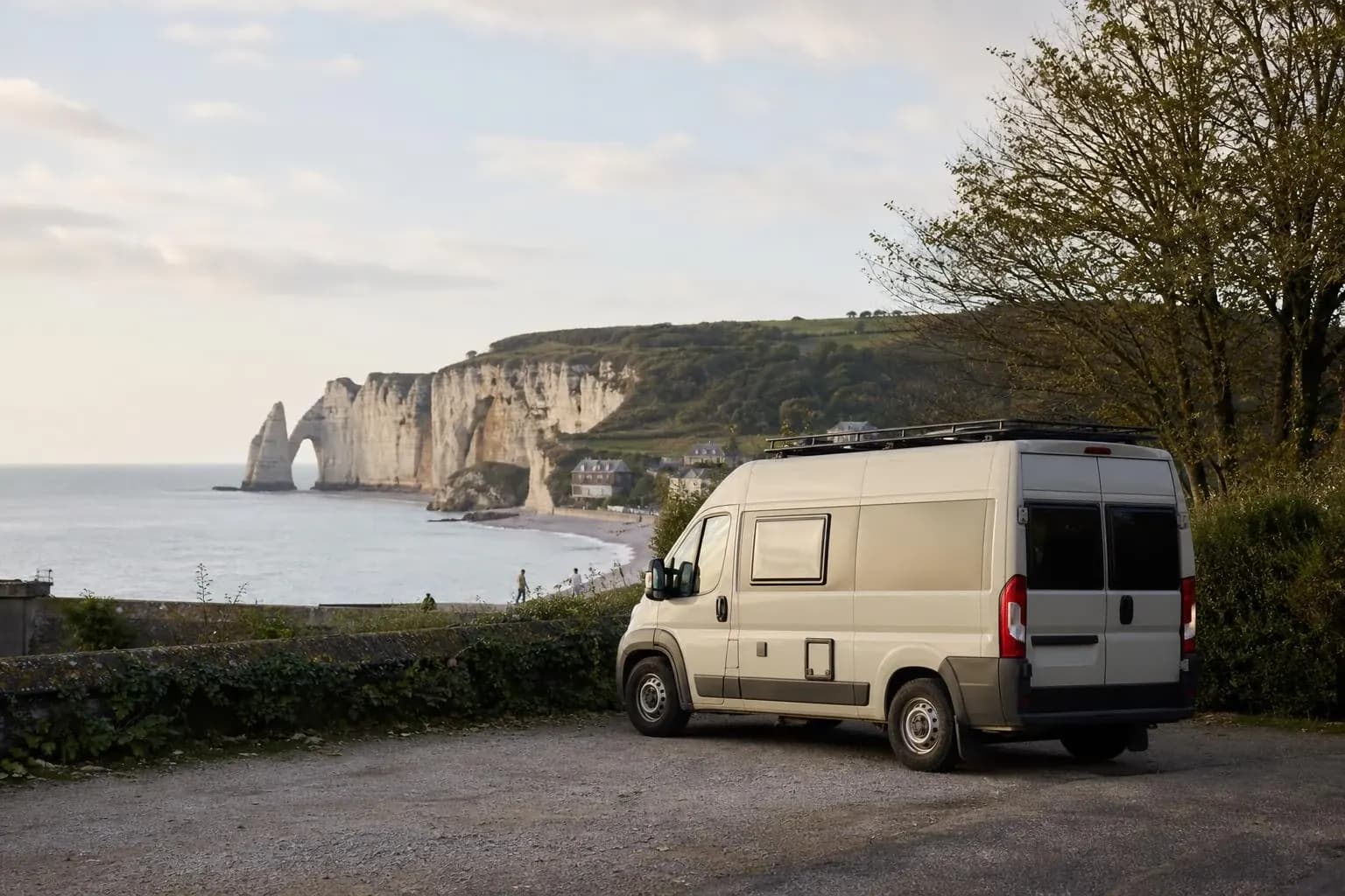Fourgon aménagé blanc-crème vu de trois-quarts arrière, garé en haut de la falaise d'Étretat au bord d'un muret de pierre, l'arche rocheuse et la plage en contrebas, lumière de fin de journée